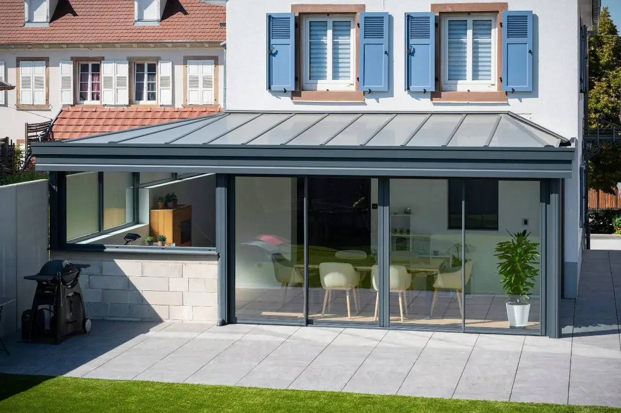 Une maison moderne avec une véranda en métal gris dotée de grandes baies vitrées, donnant sur une terrasse en dalles claires et un