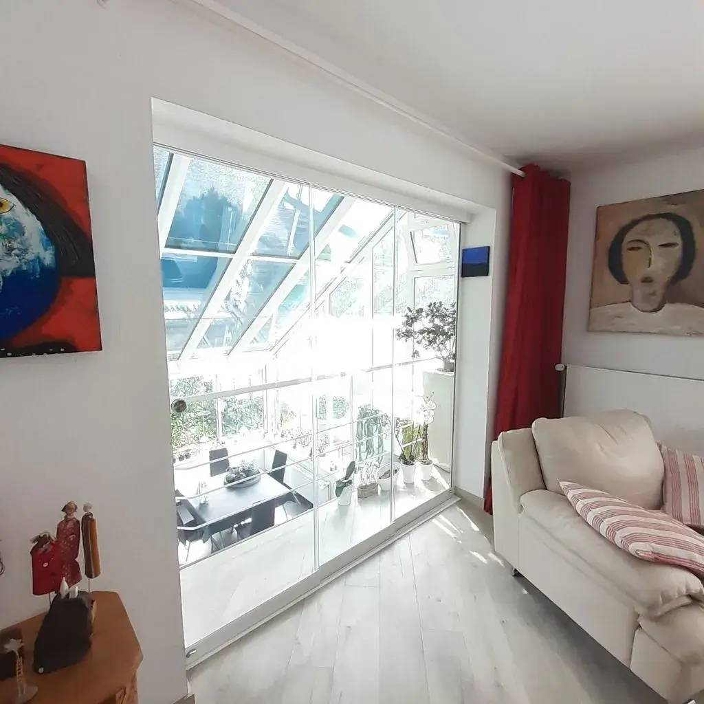 Un salon lumineux avec un canapé beige, des œuvres d'art modernes et une vue sur une véranda vitrée remplie de plantes.