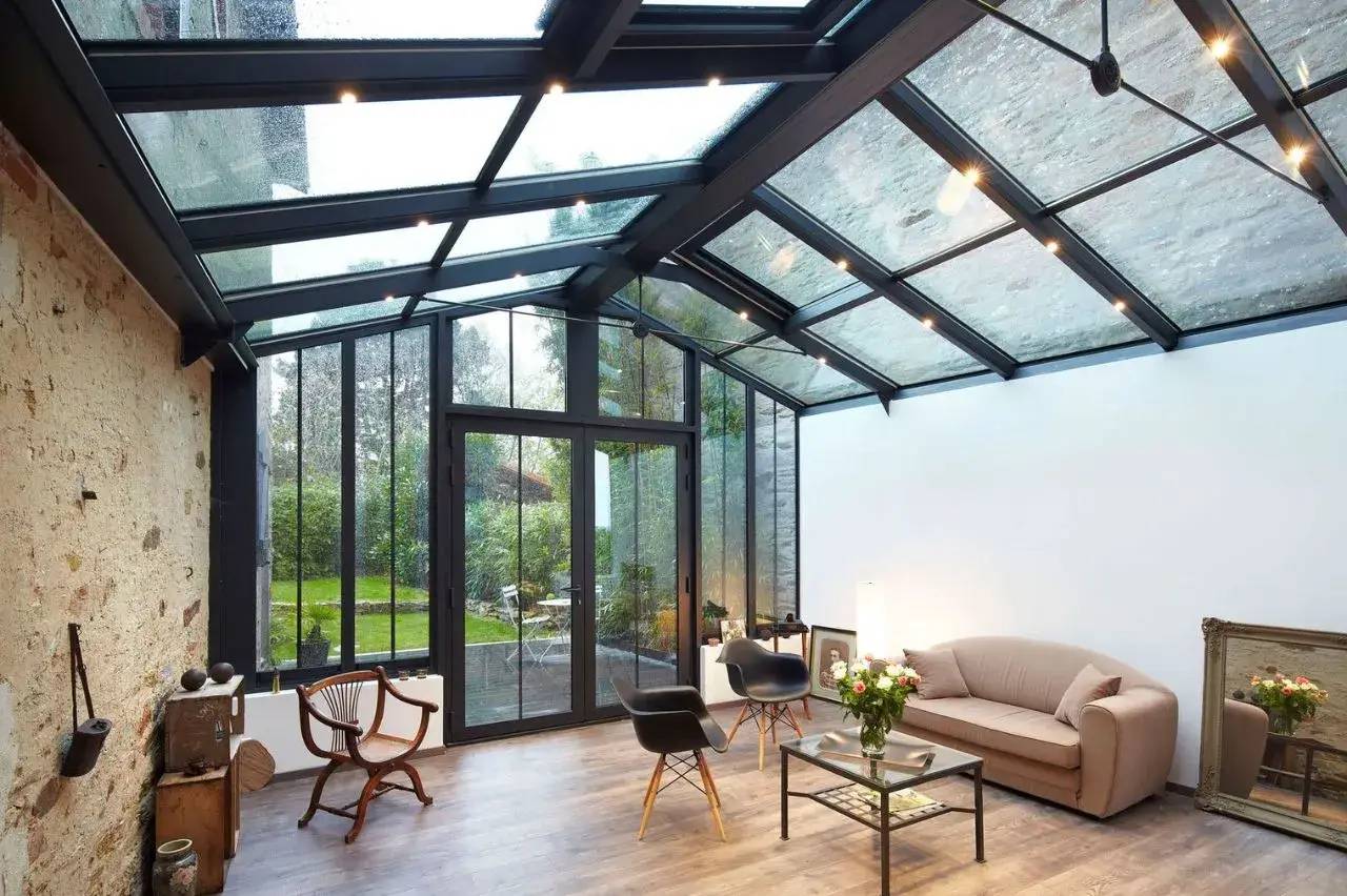 Une véranda moderne avec un toit en verre, un mobilier contemporain, et une vue sur un jardin verdoyant.