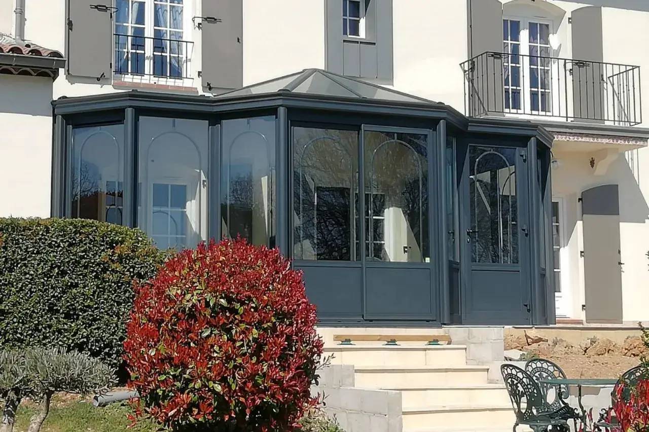 Une véranda moderne en verre et métal gris anthracite jouxte une maison avec des volets gris, entourée de buissons soigneusement
