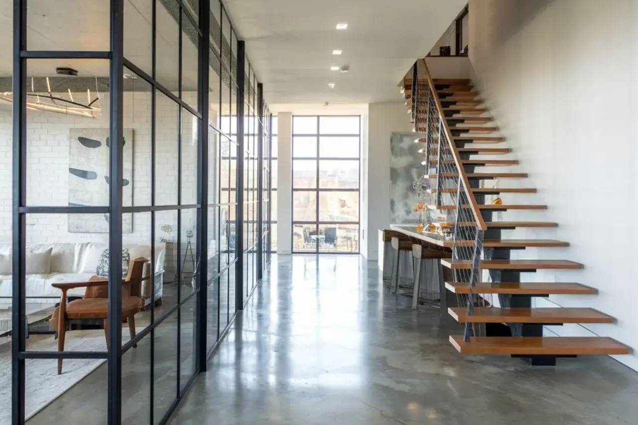 Un intérieur moderne et lumineux avec un escalier en bois flottant, des murs en verre, et un sol en béton poli.