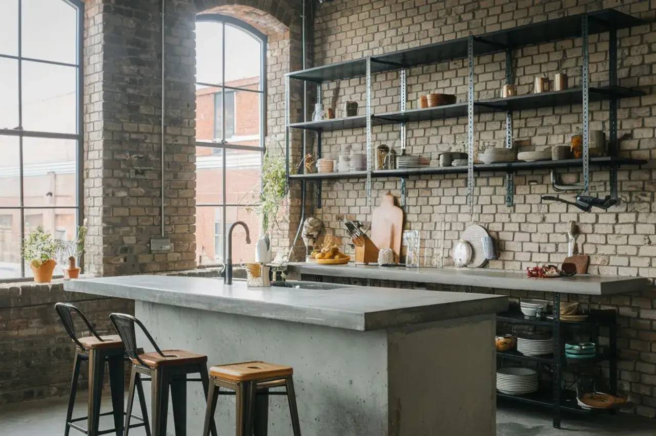 Cuisine au style industriel avec des murs en briques apparentes, un îlot central en béton, et des étagères en métal.