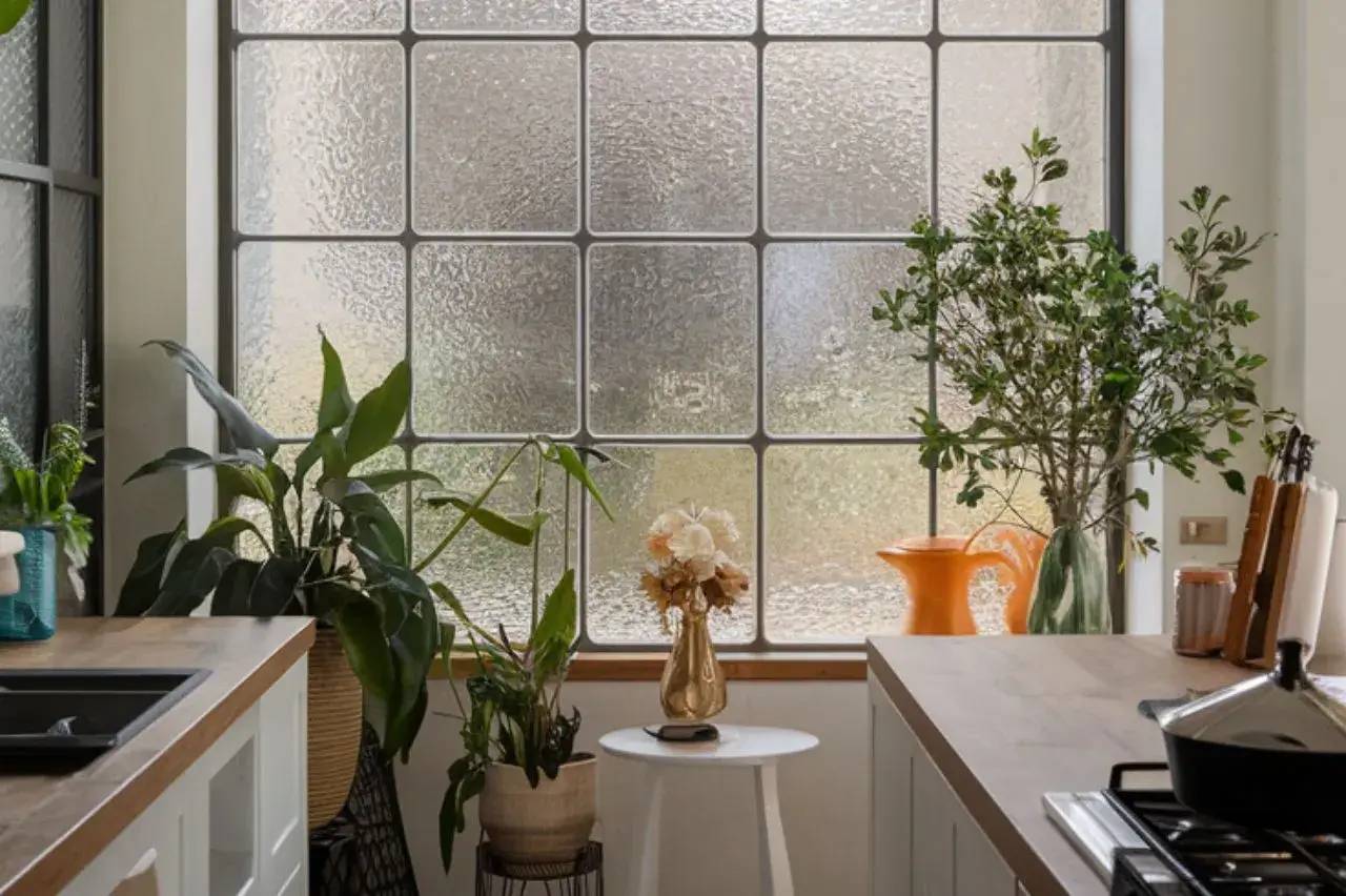 Une cuisine lumineuse avec de grandes fenêtres opaques, décorée de plantes vertes et d'un vase de fleurs sur un tabouret blanc.