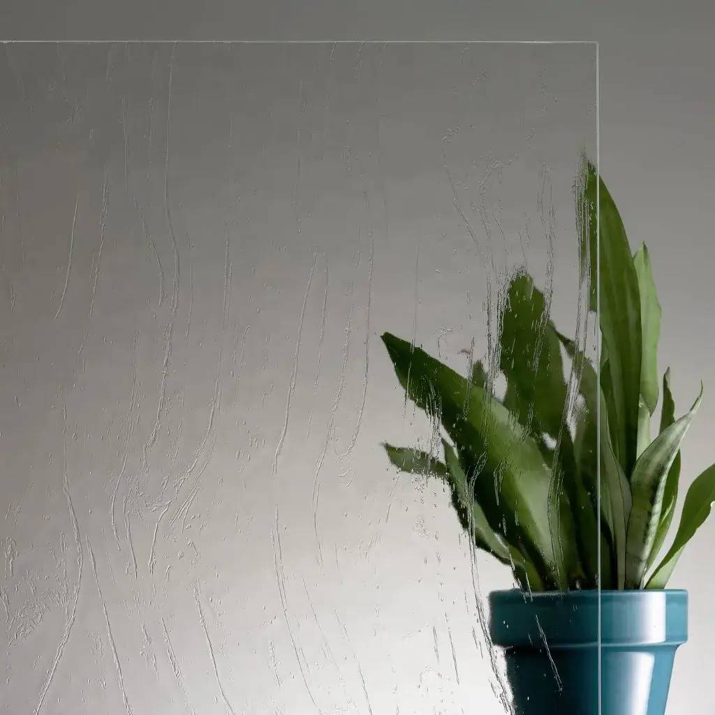 Une paroi de verre texturée devant un pot de plantes vertes sur fond gris neutre.