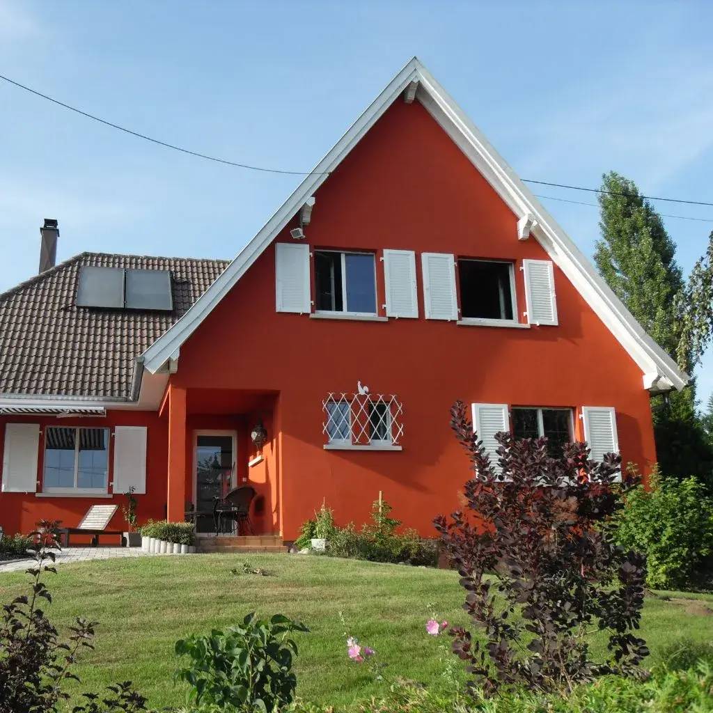 Maison rouge avec volets blancs dans un jardin, sous un ciel bleu.