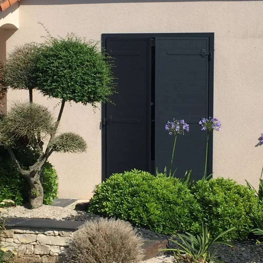 Façade de maison avec volets noirs fermés, entourée de plantes et d'un jardin bien entretenu.
