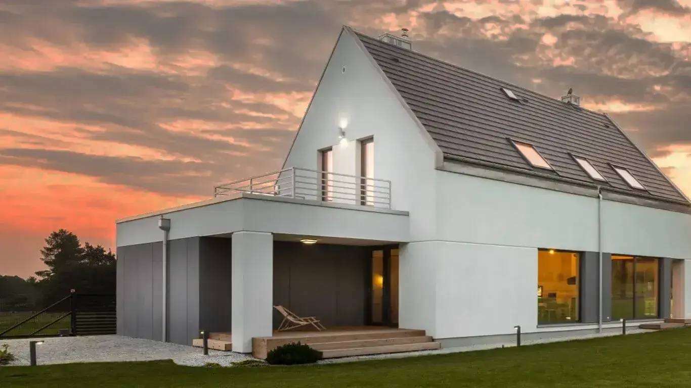 Maison moderne blanche avec un toit gris, éclairée en soirée par un ciel orangé.