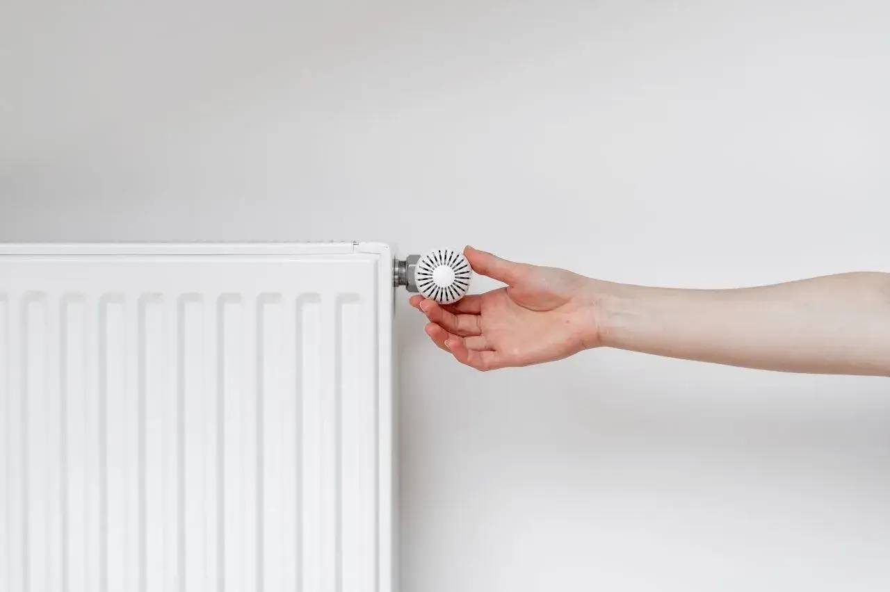 Une main tourne le thermostat d'un radiateur blanc sur un fond gris clair.
