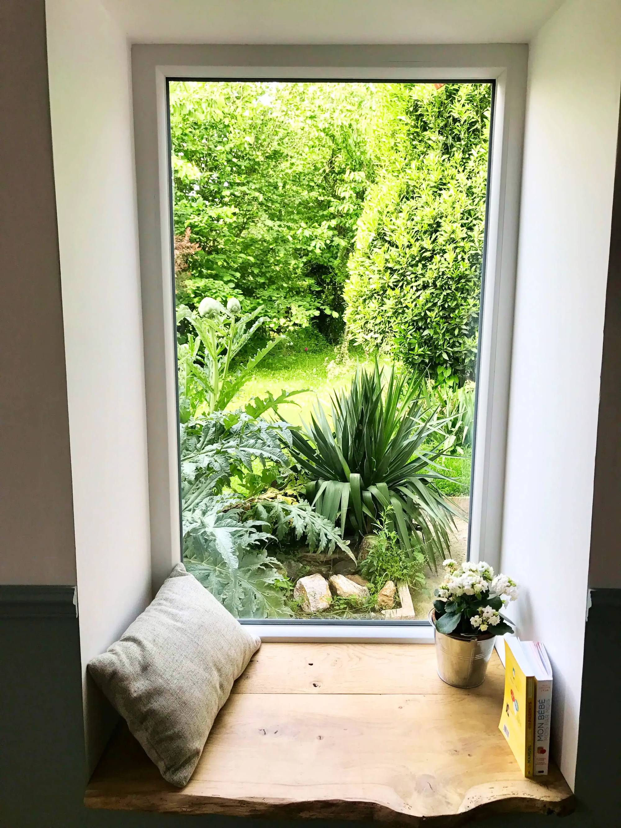 Un coin lecture cosy avec une fenêtre donnant sur un jardin verdoyant, un coussin, un livre et une plante en pot sur une étagère