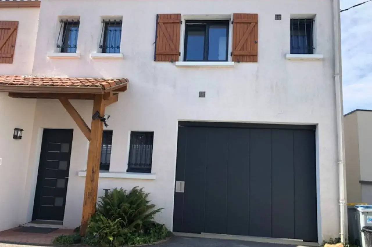 Façade de maison moderne avec porte de garage anthracite, volets en bois et petit jardin devant l'entrée.